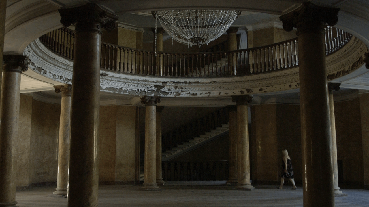 A woman is seen walking into a dilapidated large foyer area. There's a balcony above large marble pillars, and a chandelier hanging from the middle of the ceiling. Paint is peeling off the edges of the balcony, and threads of crystals are hanging off the chandelier.