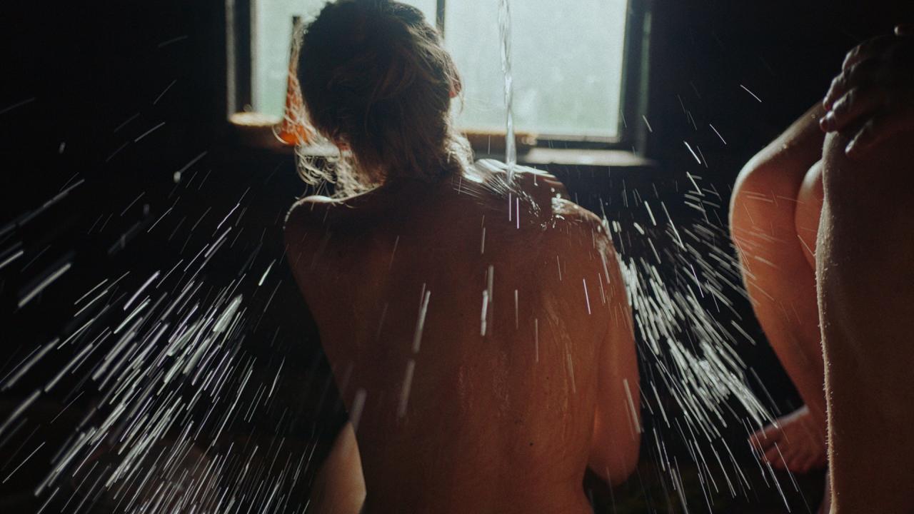 Image taken from behind of a woman in a dark room facing a window. Water is splashing against her naked back.