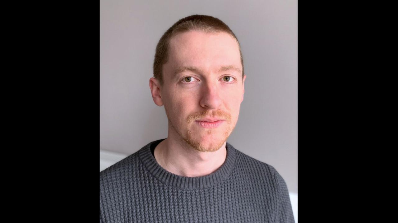 Jamie's headshot. Man with short hair is in front of a white wall. He's wearing a grey jumper. 