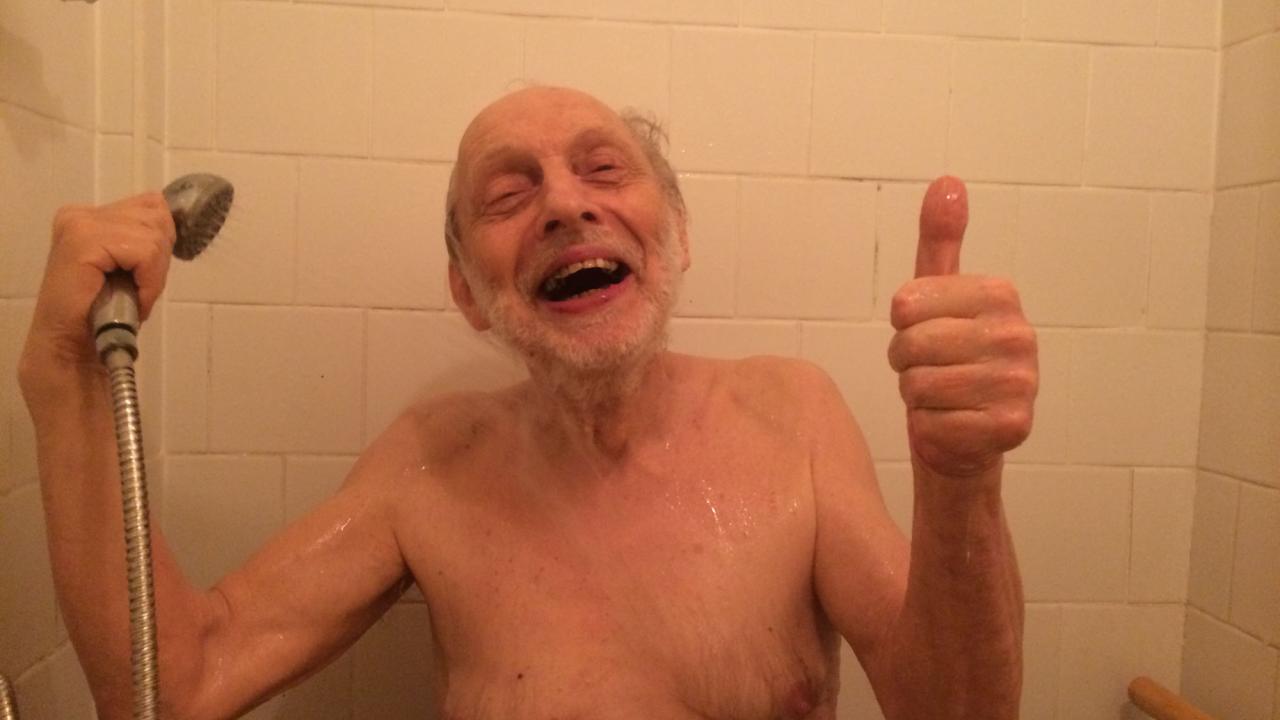 Person stands in a shower cubicle, in one hand holding the shower head and with the other hand putting his thumb up 