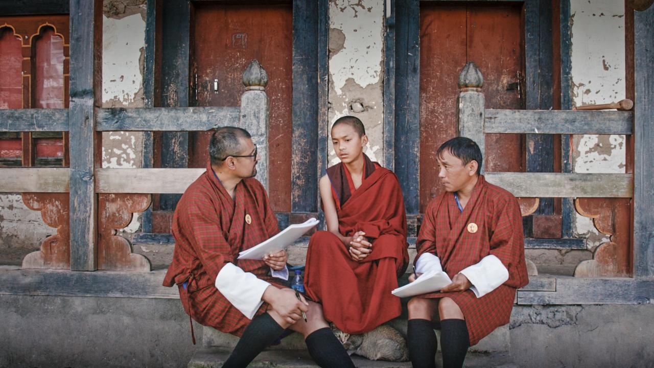 Three people sat in traditional dress sat on steps. The person on the left and the person on the right hold a stack of paper each.