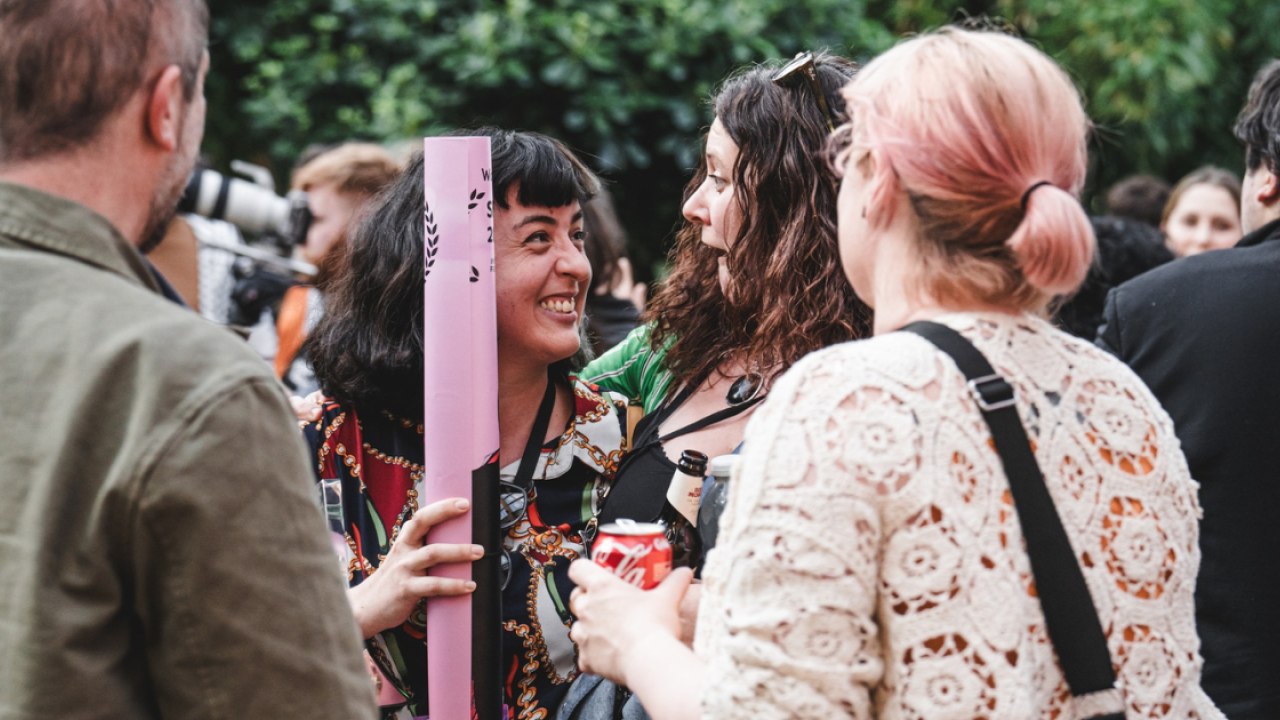 Two people looking at each other smiling widely, surrounded by green foliage. The person on the left holds a rolled up film poster in their hand.