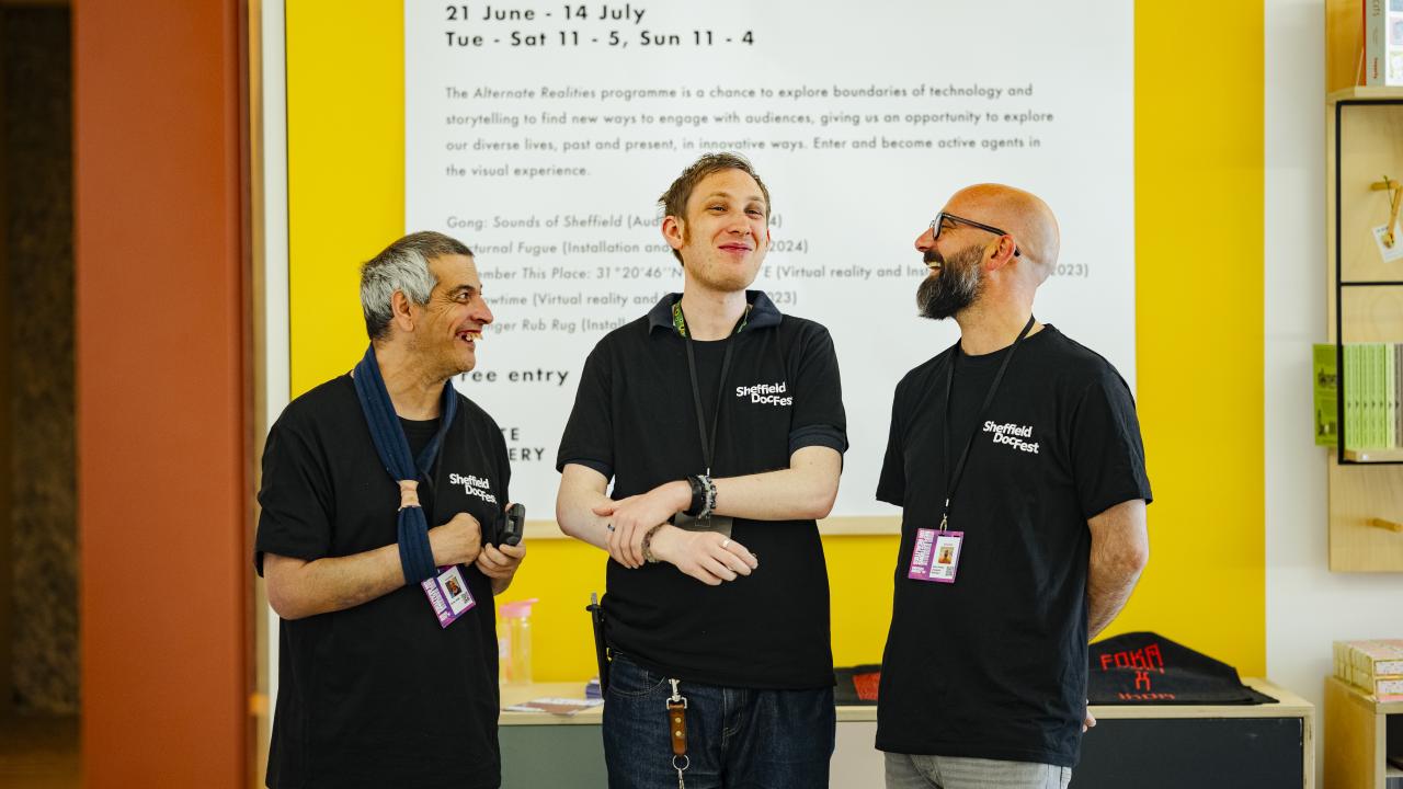 Three people stand in a line but turned towards each other. They are wearing t-shirts with the Sheffield DocFest logo and are smiling widely.