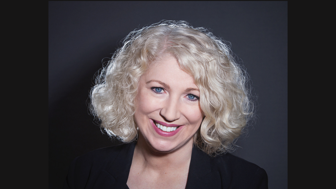 Headshot of a woman with blonde, chin length, curly hair. She is wearing a black blazer and is pictured against a dark grey backdrop.