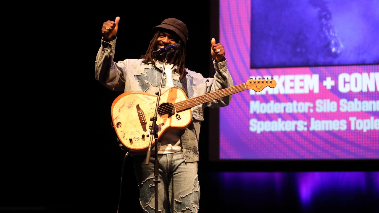 Man stood on stage wearing a guitar with both thumbs up.