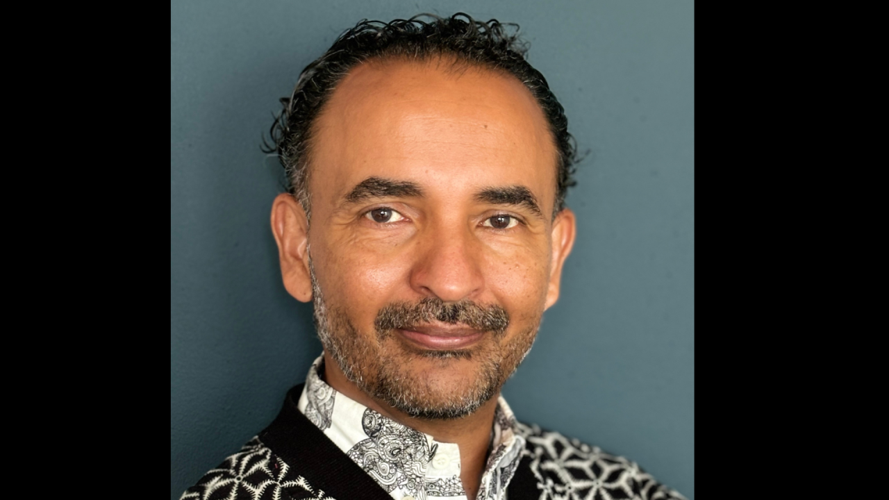 Raul Niño Zambrano headshot. Image of a man with dark curly hair and facial hair. He is wearing a white shirt with a black and white patterned jumper over the top.