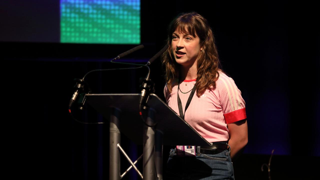 A woman in a pink top stands at a lectern in a dark room, with a spotlight on her