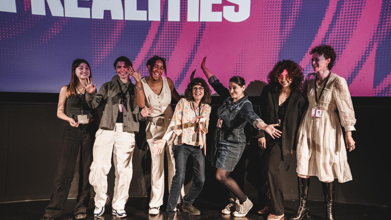 A group of people stand on a stage smiling widely with their hands in the air.