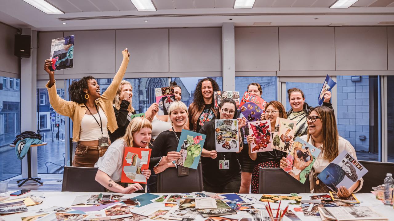 A group of people with wide smiles and their hands in the air holding collages they have made.