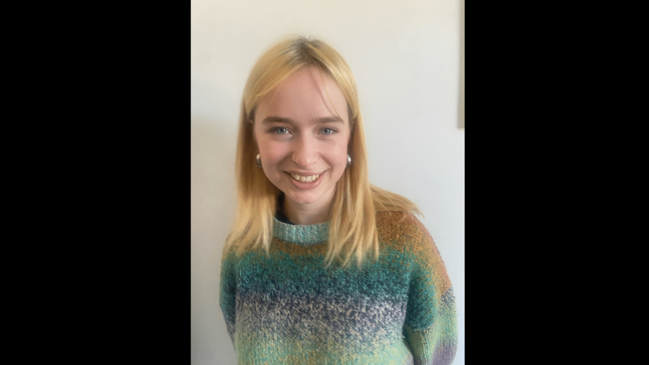 Phoebe Hickson headshot. Image of a white woman with straight blonde hair and blue eyes. She is wearing a colorful jumper and is smiling. She is against a white wall.