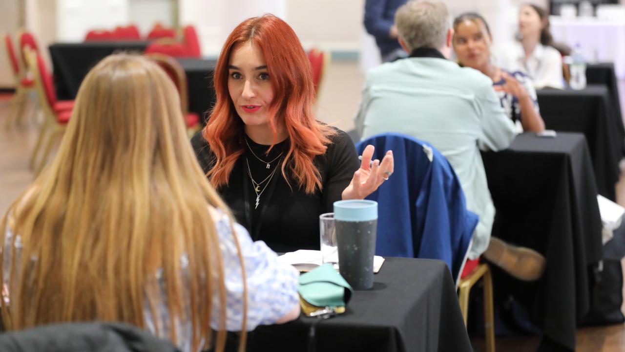 A line of tables with people either side of each table chatting animatedly. The most prominent person in the image is a person in the middle of the photo with long red hair. 