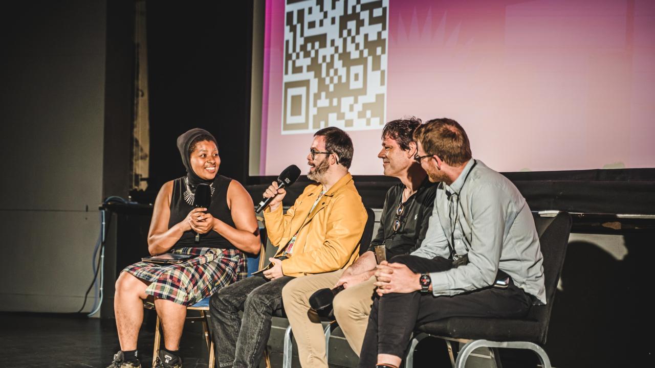 A group of people sat on a stage chatting to each other holding microphones. Behind them, a large pink screening with a QR code.