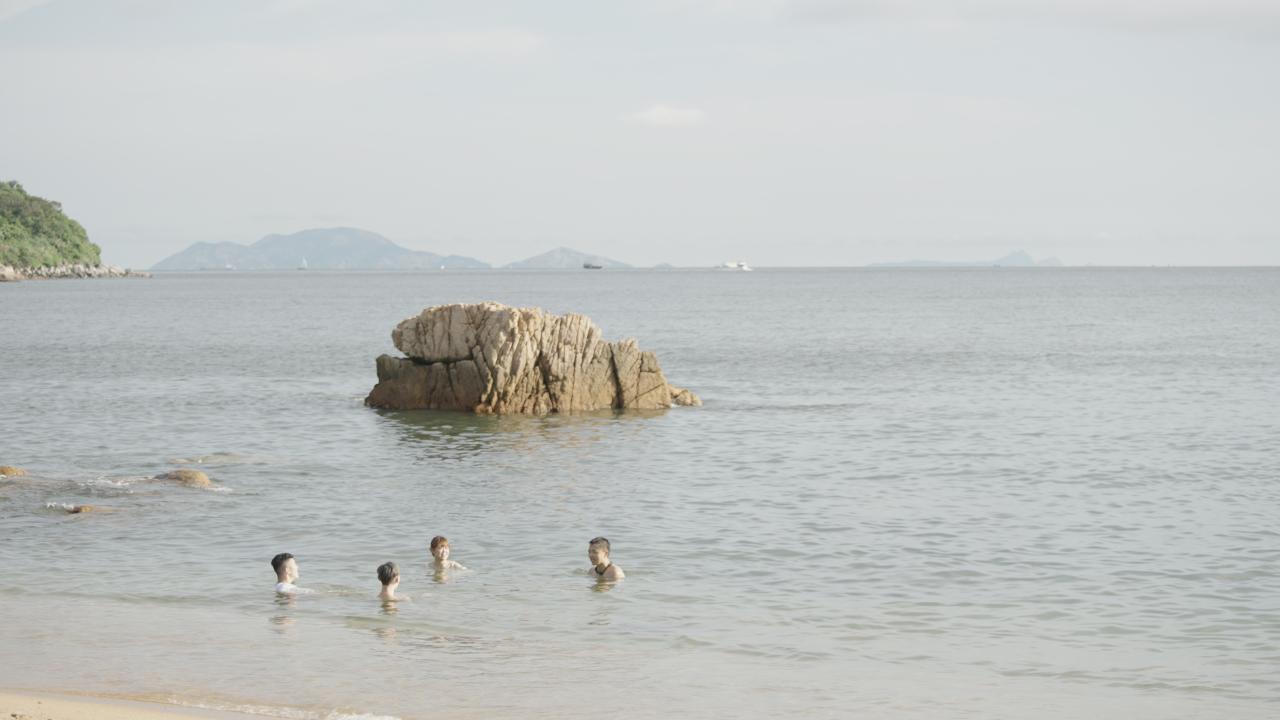 Four people swimming near the shore with a large rock formation in the sea behind them and distant mountains on the horizon.