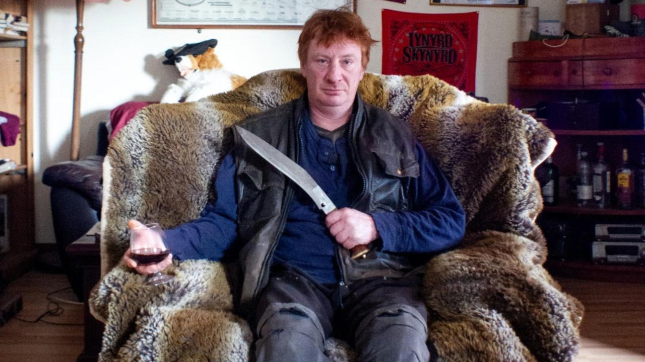 Man with red hair sitting in fur coverded chair with a knife in his left hand and a glass of wine in his right hand