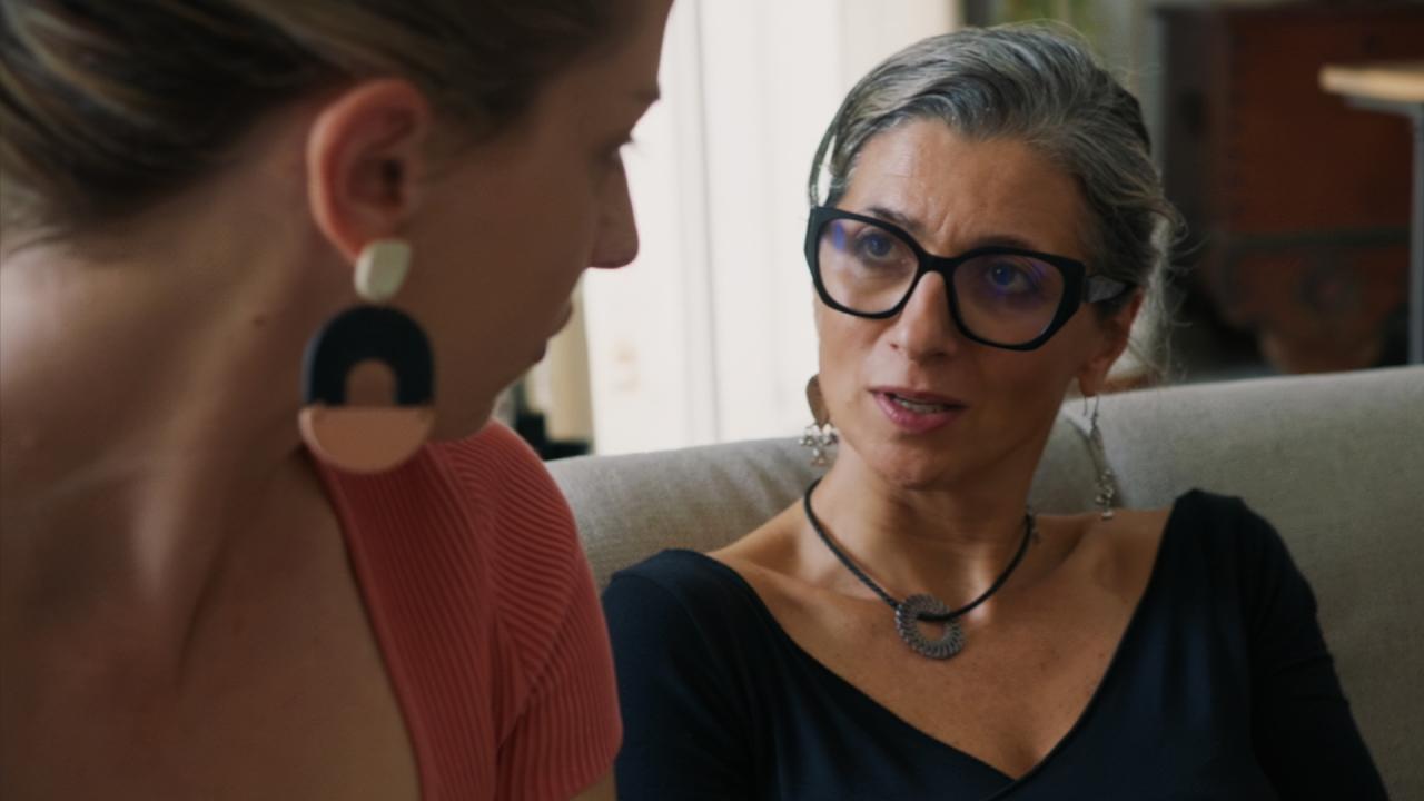 Two women sitting close together in conversation, the one more prominent in the picture with grey hair and large black glasses.
