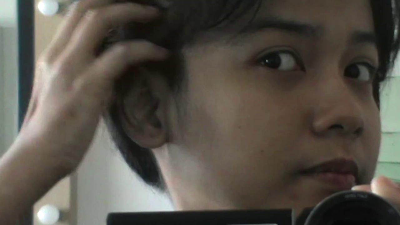Close-up of a person looking into a mirror while adjusting their hair, with a camera partially visible at the bottom of the frame.
