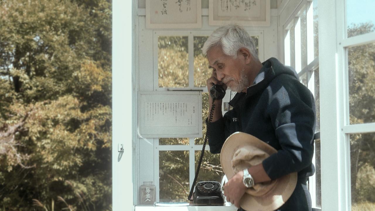 An elderly man with gray hair and a beard stands inside a white phone booth, holding a rotary phone to his ear and a straw hat in his other hand, surrounded by leafy green trees.