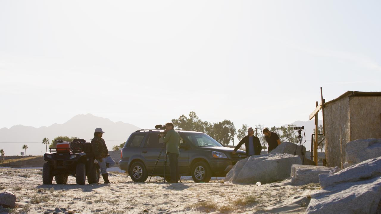A small film crew sets up beside a car in a desert landscape, with mountains in the distance and sunlight casting long shadows.