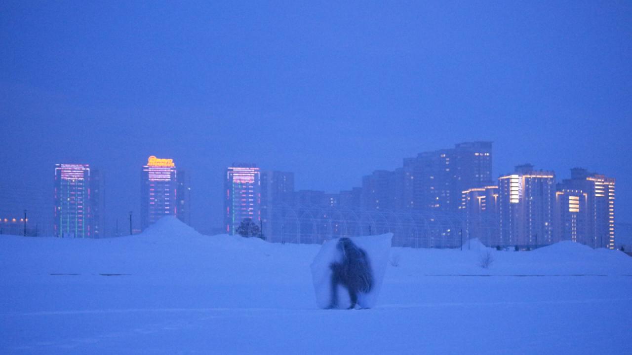 A figure crouches beneath a transparent sheet in a snowy field at dusk, with illuminated high-rise buildings glowing in the distance.