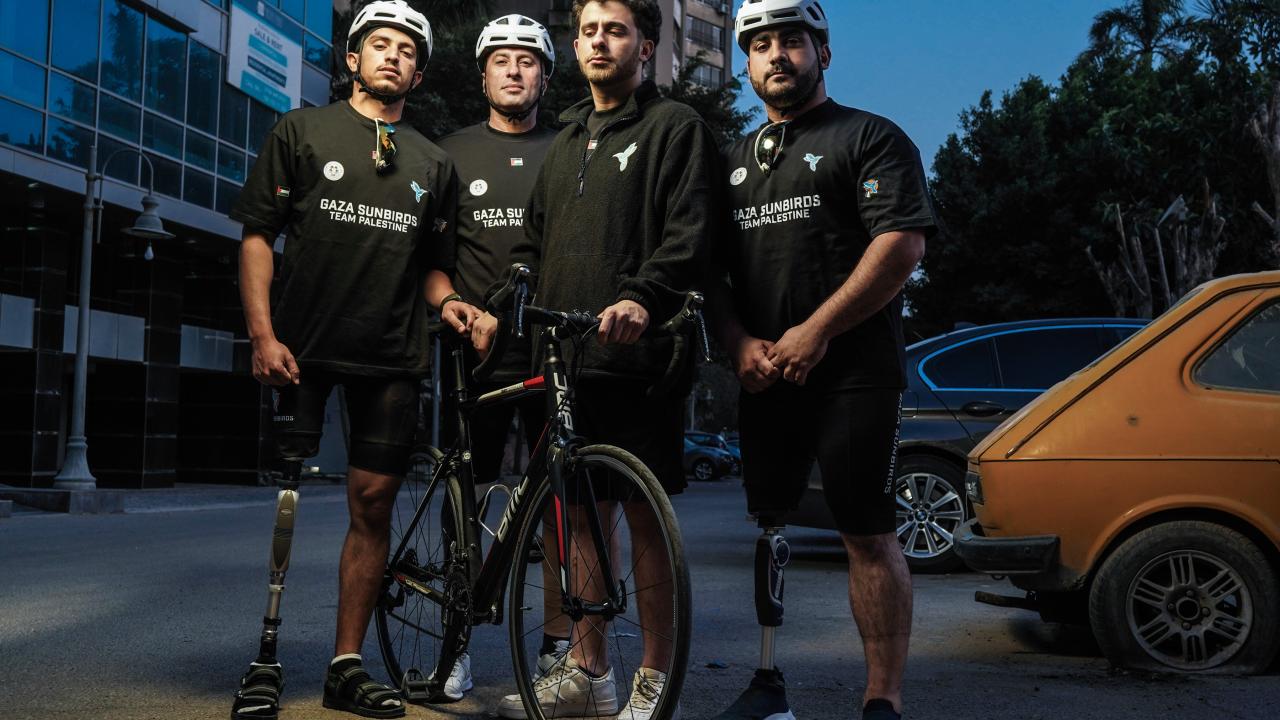 Four members of a team stand confidently with a bicycle in an urban street setting at dusk. Two of them wearing prosthetic legs, all dressed in team black shirts and three wearign helmets, illuminated by dramatic lighting.