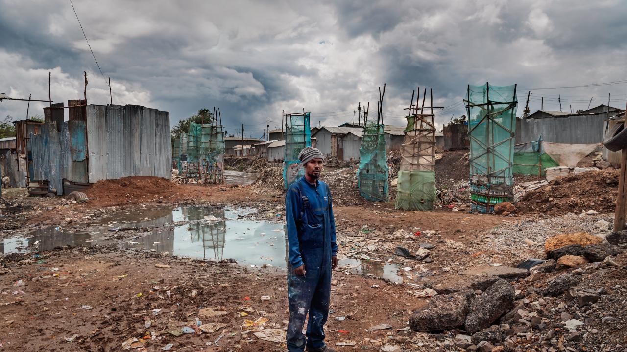 A black man in a paint-stained blue jumpsuit and striped beanie stands amid a muddy, debris-strewn construction site. Behind him, skeletal building frames covered in green netting rise against a backdrop of dark, stormy clouds.