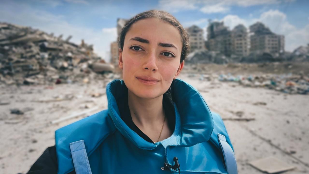 A woman wearing a bright blue protective vest stands in the foreground, looking directly at the camera. Behind her is a scene of destruction: large piles of rubble and partially collapsed buildings under a partly cloudy sky. The setting appears to be a disaster or conflict zone.