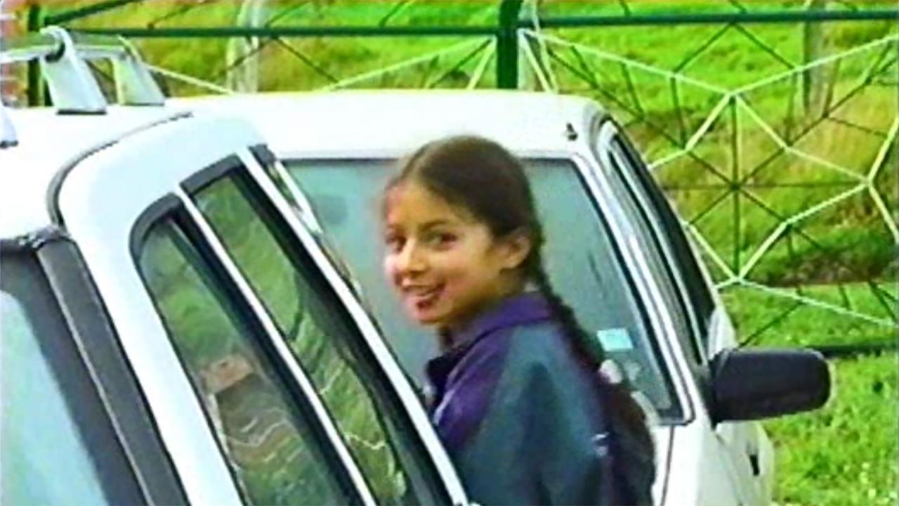 A young girl with braided hair and wearing a dark jacket smiles while standing between two white vehicles. Behind her is a green field enclosed by a fence with a geometric pattern. The image has a grainy, VHS-like quality.