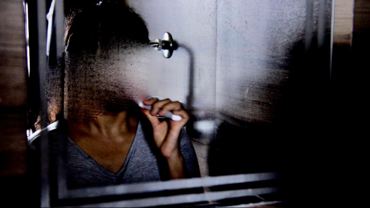 A person in a grey shirt is brushing their teeth in front of a fogged-up mirror. The mirror is partially wiped, but their face is still obscured by condensation and darkness.