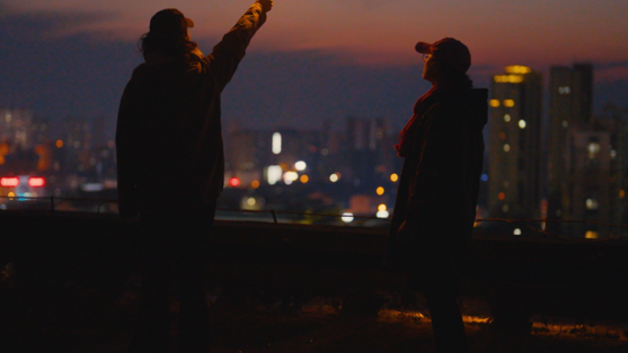 Two figures standing on a rooftop at dusk or early night, with a cityscape glowing in the background. One person is holding a lit sparkler aloft, casting a warm light and small sparks into the air. The silhouettes of both individuals are visible against the fading light of the sky.