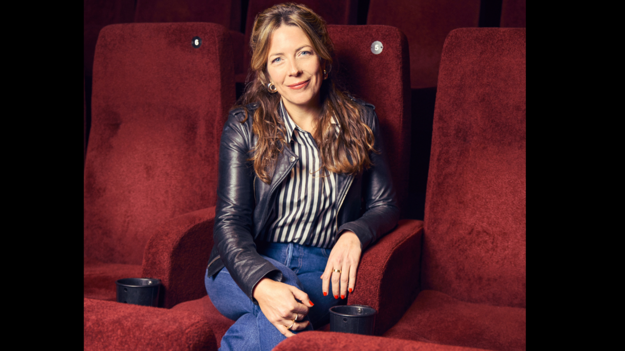 Photo of Mimi headshot. She is sat on red chairs, wearing a leather jacket with denim jeans. She has long dark blonde hair, half up, with blue eyes. She looks casual. She is smiling. 