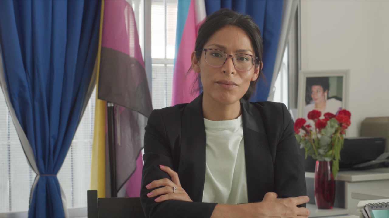 A composed woman with black hair and glasses sits confidently at a desk, arms crossed, her expression serious yet calm. Behind her, pride flags and rich blue curtains frame the setting, along with a framed photo of another woman and a vase of red flowers.