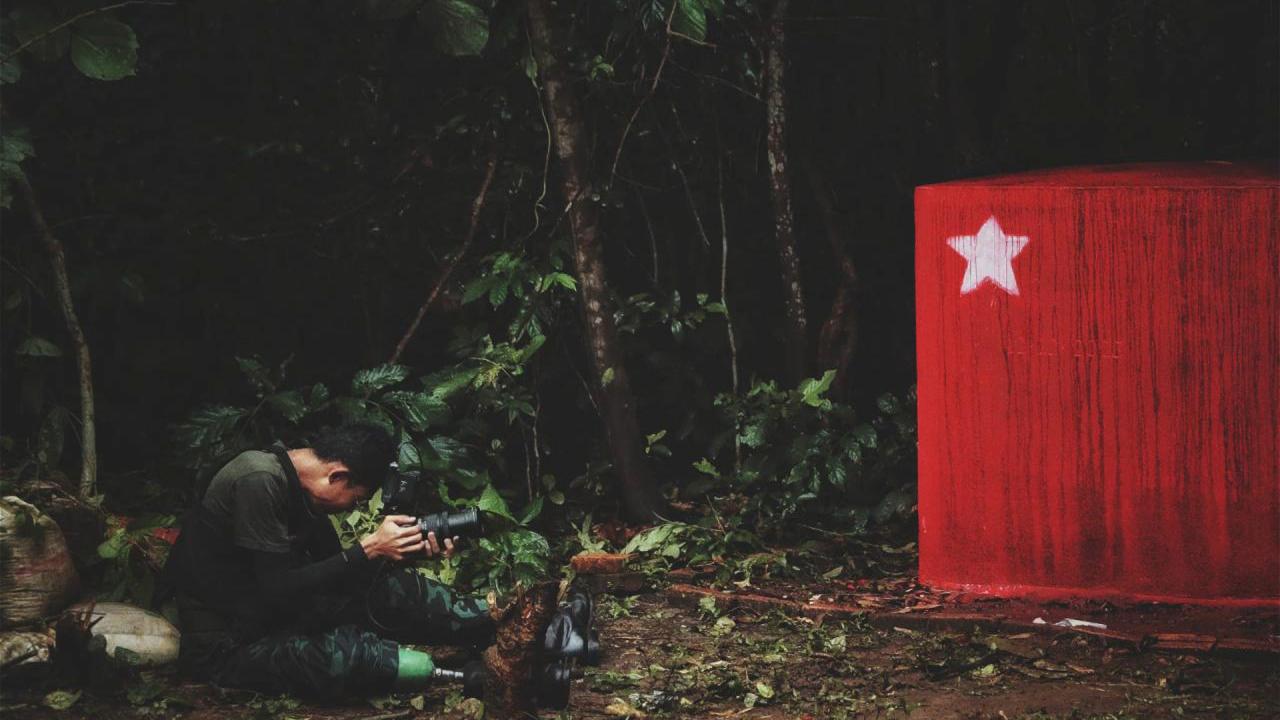 In a dim, dense forest, a man in camouflage gear crouches low, focusing intently through the lens of his camera. Beside him and on the right side, a bold red monument with a white star stands strikingly against the greenery.