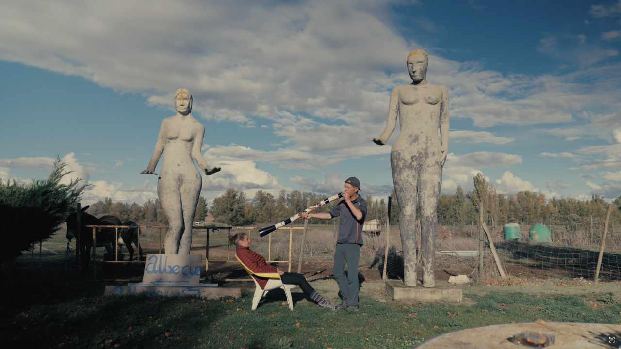 A man dressed in blue with a blue hat plays a long, cylindrical wind instrument decorated with black, white, and red geometric patterns toward a seated woman with a red jacket between two large, weathered statues in a rural field.