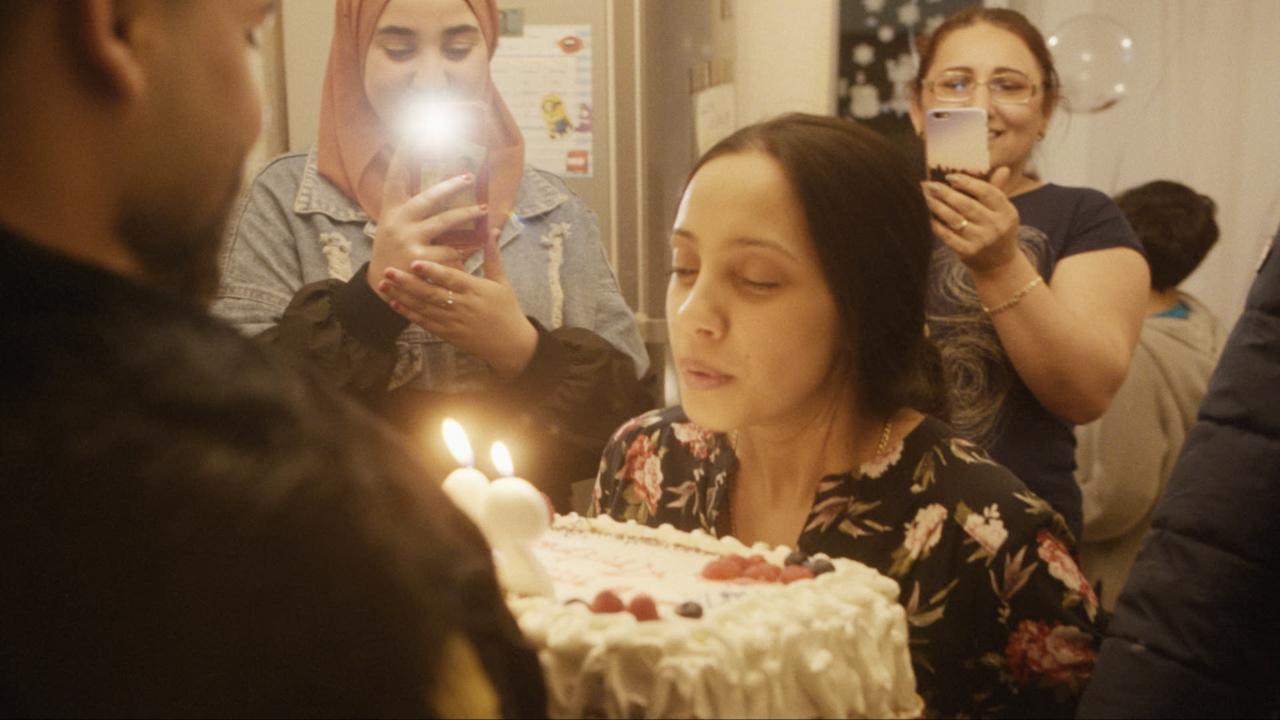 A woman in a floral dress is about to blow out two lit candles on a birthday cake, while others around her, smiling and holding up phones, capture the moment in a warm indoor lighting and joyful setting.