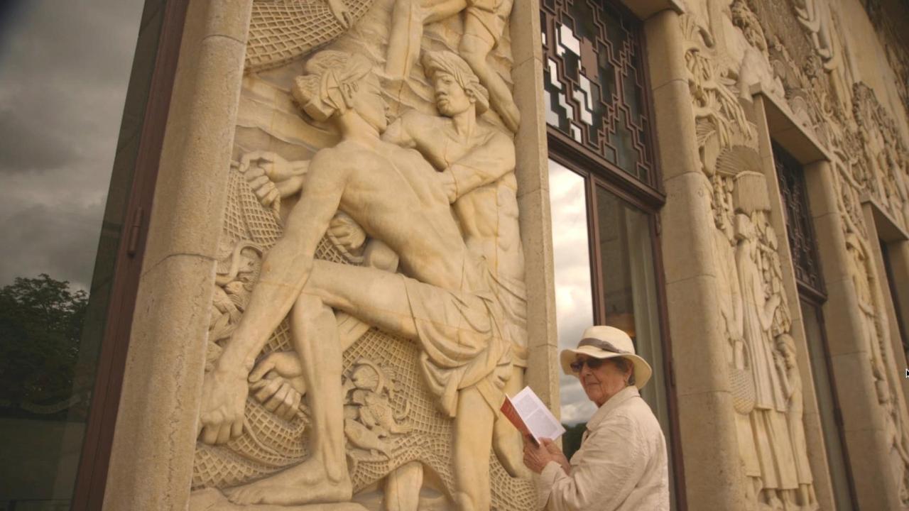 An older woman in a sunhat and light jacket stands reading from a booklet in front of a grand stone wall. The beige wall is intricately carved with muscular human figures. 