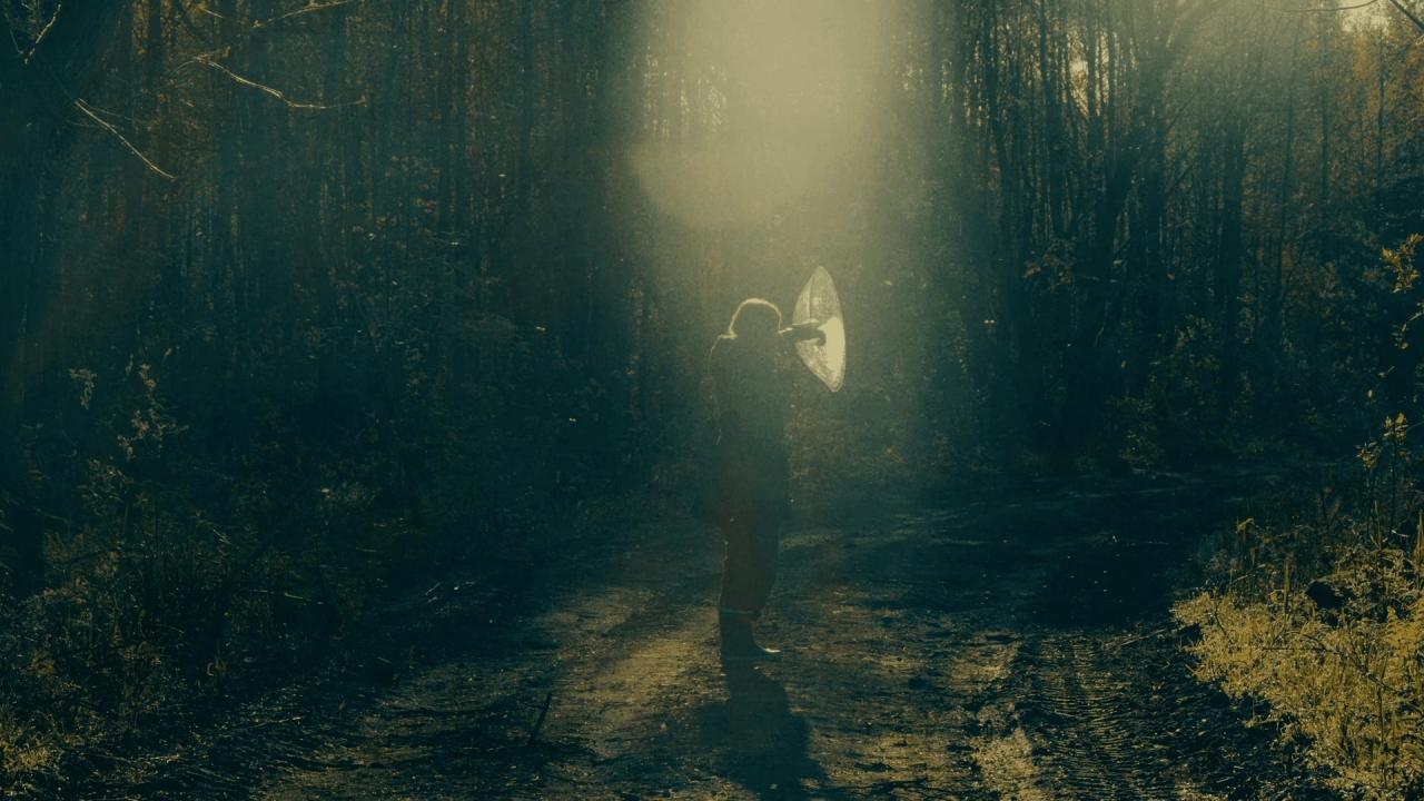 A lone figure stands on a dirt path in a dark, shadowy forest, bathed in a beam of ethereal light. They hold a large translucent disc.