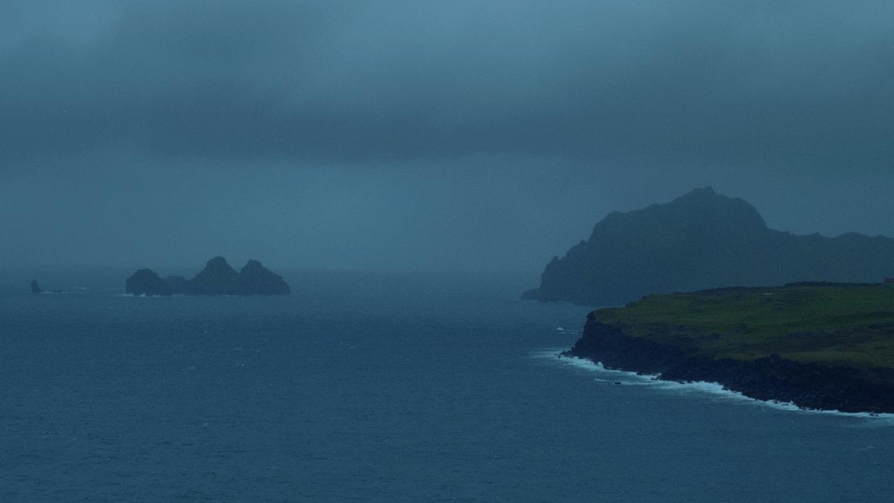 A brooding coastal scene under overcast skies, where stark cliffs stretch into the sea from the right, their dark rock faces contrasting with a narrow strip of green grass above. In the distance, rugged islets rise from the water, shrouded in mist and shadow. 