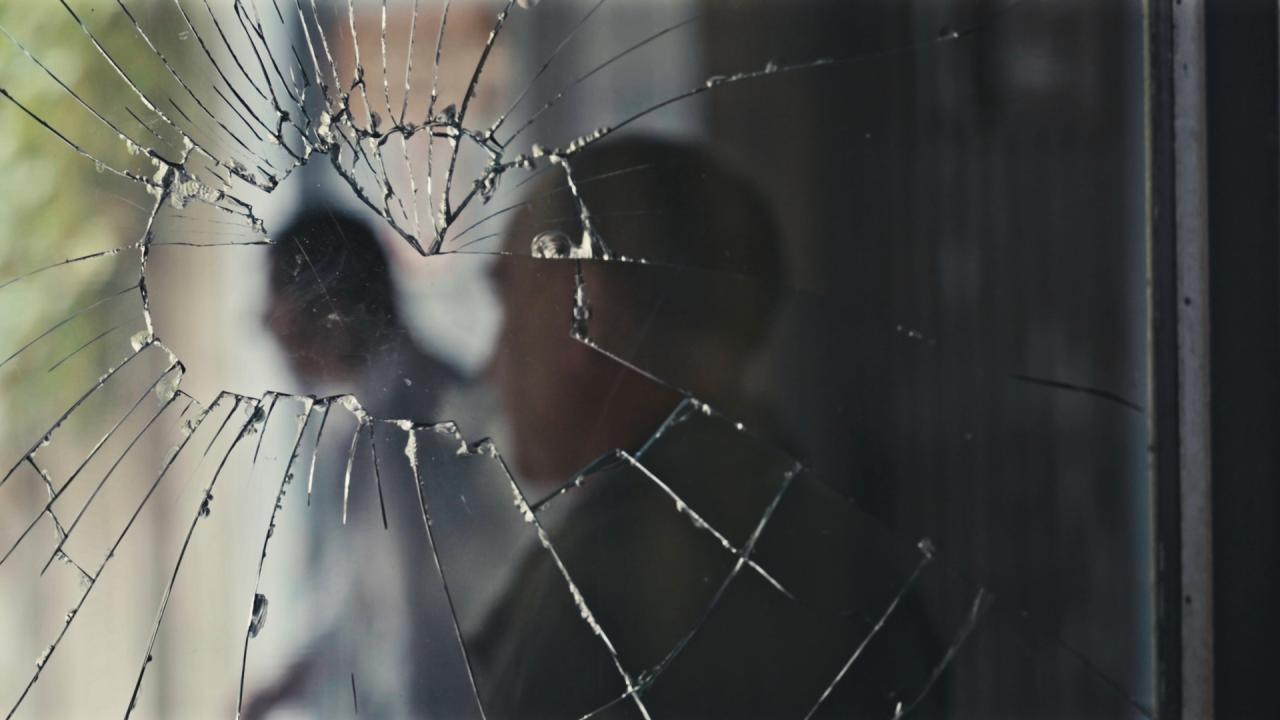 A cracked windowpane through which we glimpse a blurred human figure in the background. The broken glass radiates outward from a central impact point, creating a spiderweb-like pattern that visually fragments the person beyond.