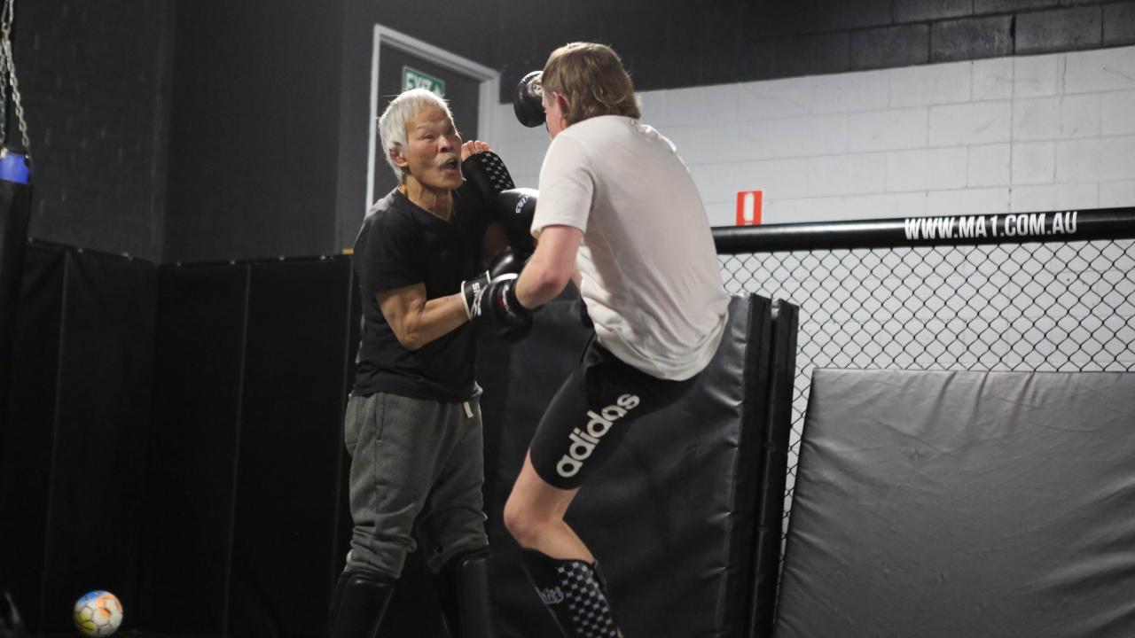 Two people are sparring or training in a martial arts gym. One older Asian person with grey hair is holding focus mitts while the younger person in gloves and shin guards is practicing strikes. They are inside a caged training area with padded walls and a soccer ball in the corner.
