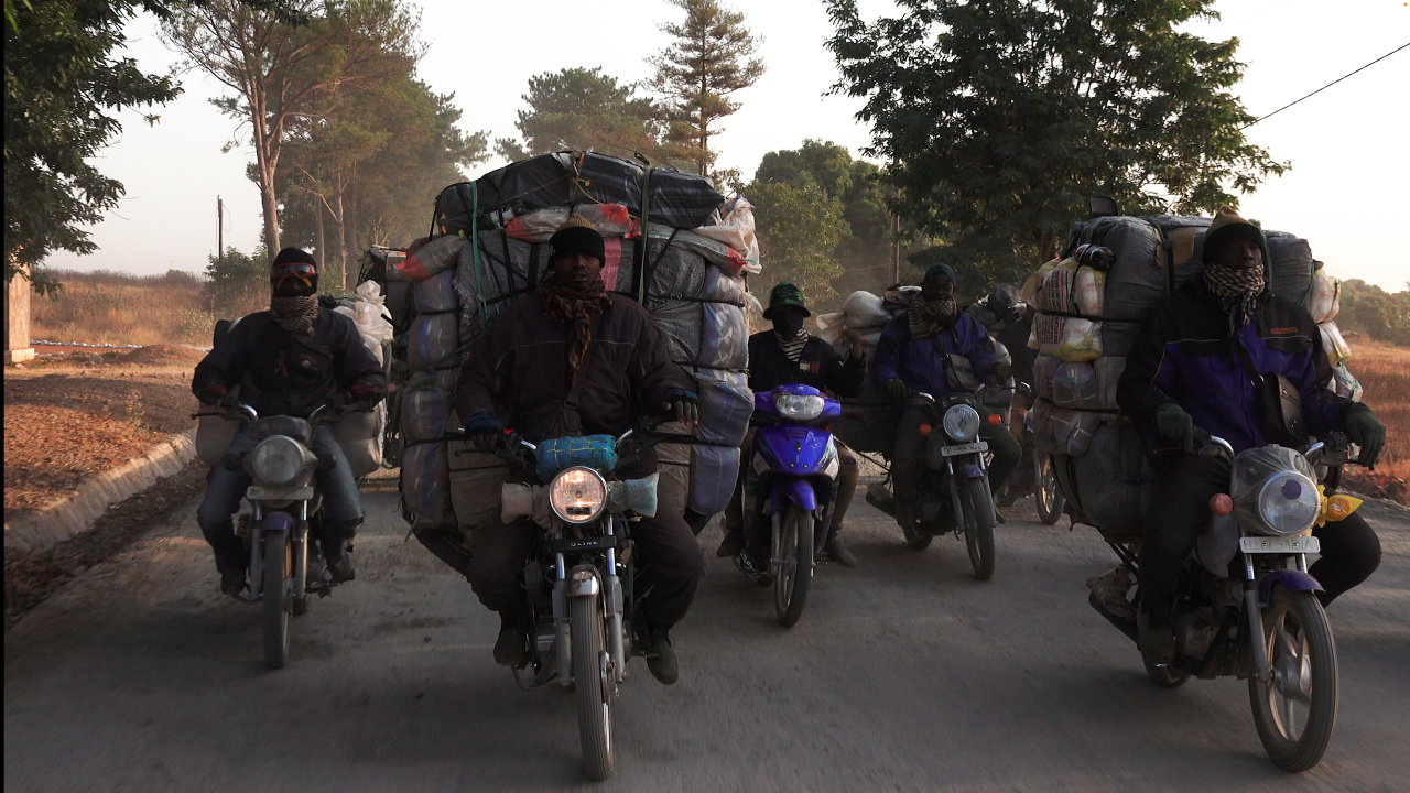 A group of men riding motorcycles heavily loaded with large, bundled cargo. There are six visible riders, all wearing dark clothing, gloves, scarves or masks covering their faces, and hats or helmets. The motorcycles are driving on a paved rural road flanked by dry vegetation and trees. 