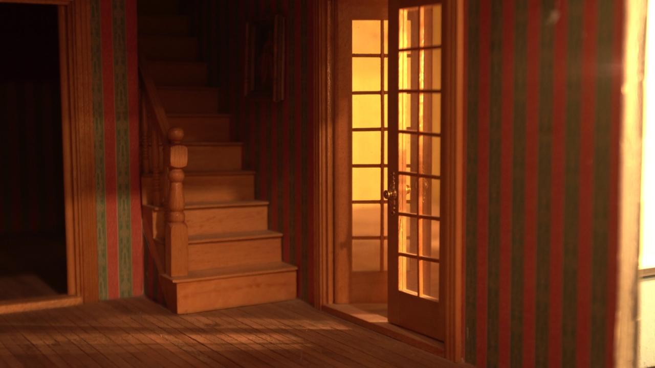 A small, dimly lit interior with warm lighting coming from a room beyond a set of glass-paneled doors. There is a staircase leading upward to the left and wooden flooring throughout. The wallpaper is vertically striped in red and green. 