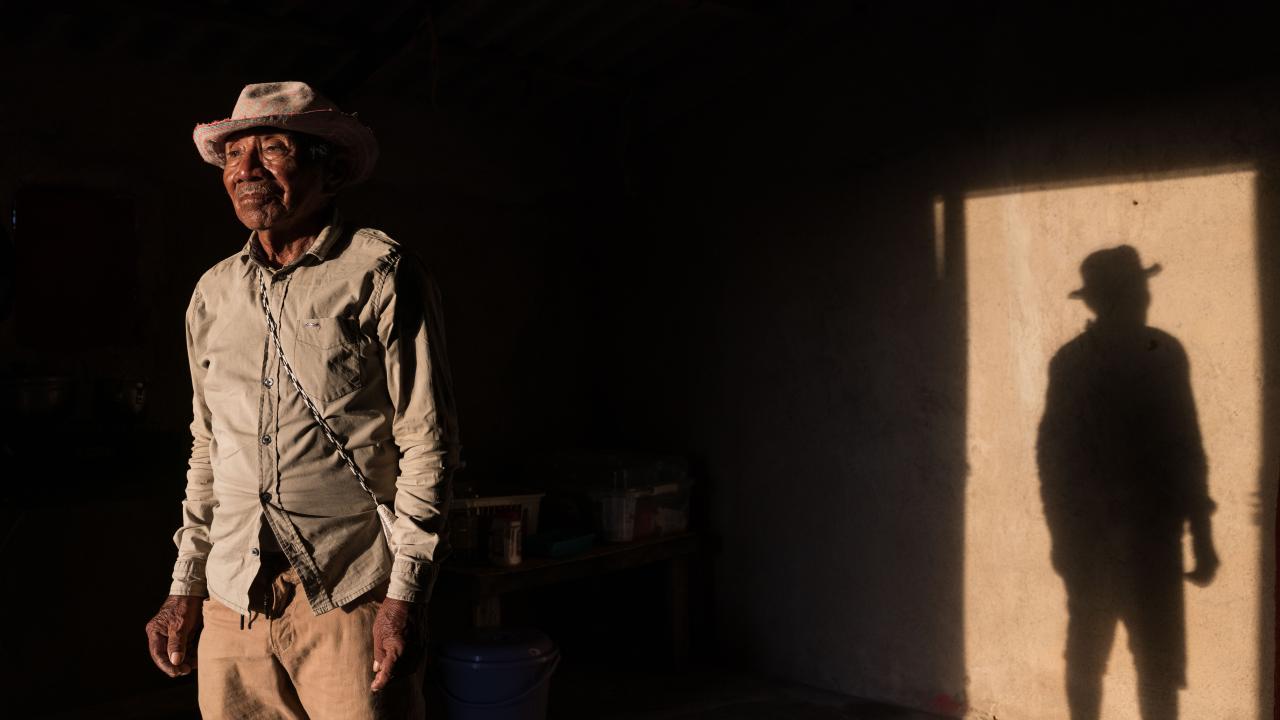 An older man standing in a dimly lit room, with strong light casting a long, defined shadow of him on the wall. He wears a hat and simple clothing, with a contemplative or slightly solemn expression. 