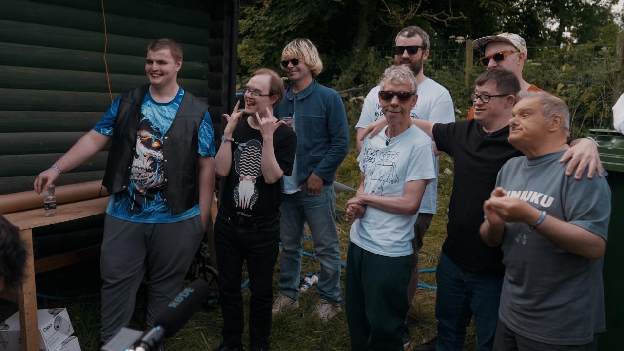 A group of people, likely members of a community or group, enjoying a moment together outdoors. The expressions are joyful, with many of them smiling or making hand gestures that show togetherness and camaraderie. They are looking towards the left of the picture.