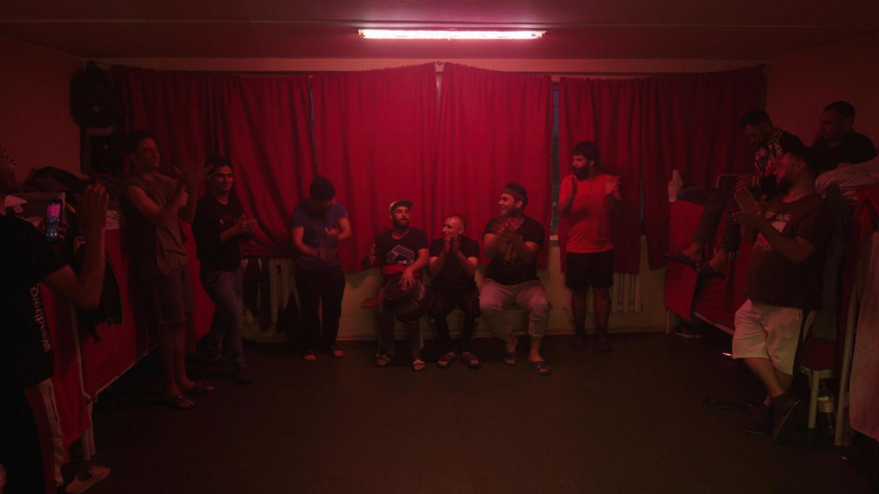 A group of men gathered in a dimly lit room with red neon lighting, clapping and playing music, some seated and others standing.