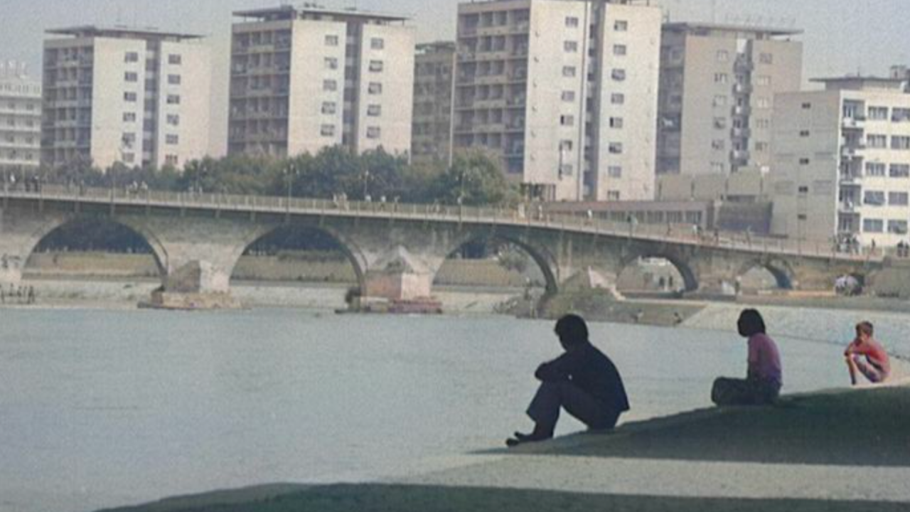 In a quiet, contemplative atmosphere, three figures sit in the foreground on the right of the screen along a riverbank, their poses subdued and introspective. Behind them, a stone bridge connects two urban zones, its arches mirrored in the calm water. 