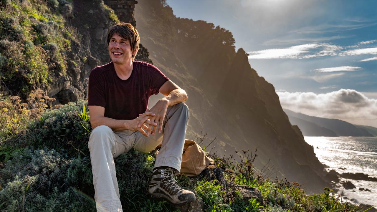 Brain Cox sat on a large rock with a sunny hillside behind him. On the far right the sun shines on the ocean