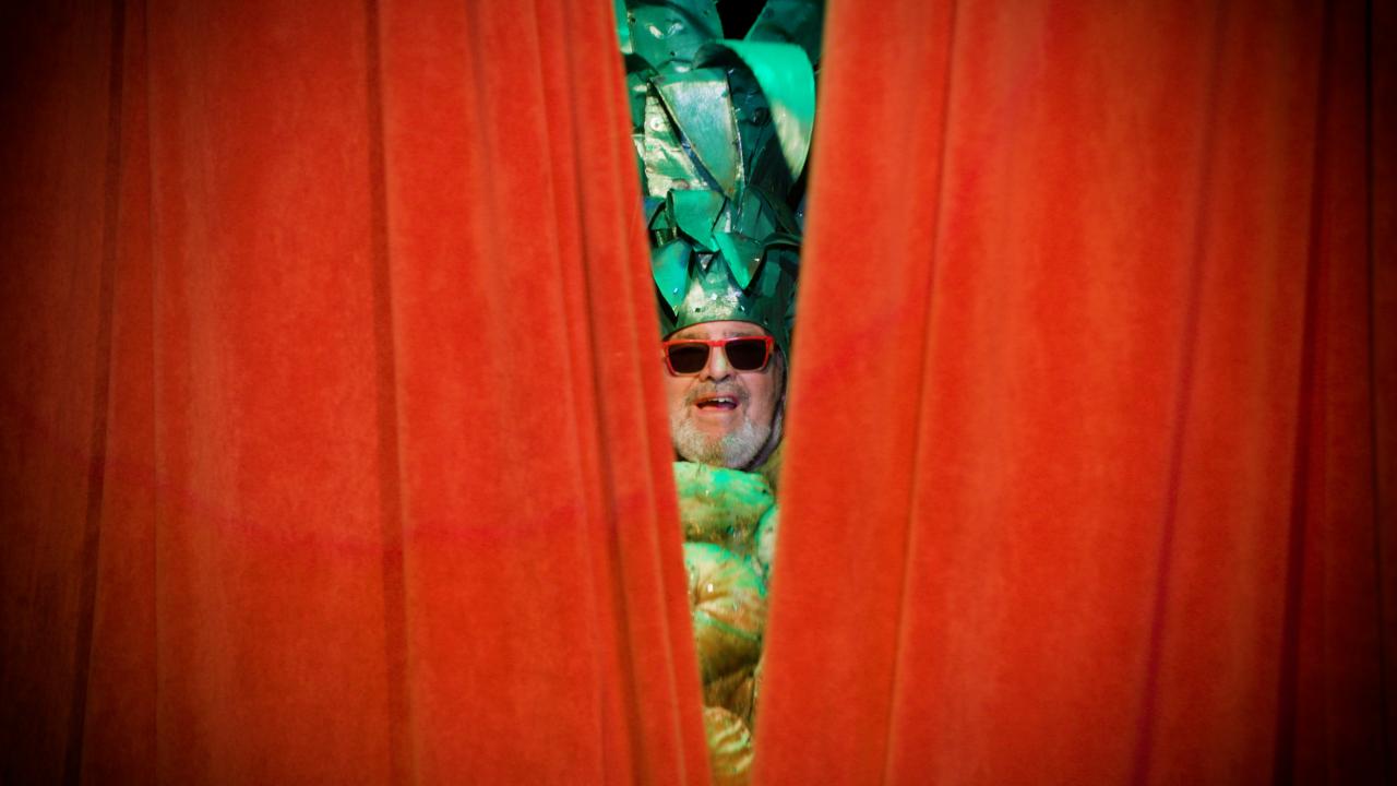 Large red theatre curtains cover most of the image, except for the very center where a man's face peeks through. The man is wearing a pineapple costume and a pair of large red sunglasses.