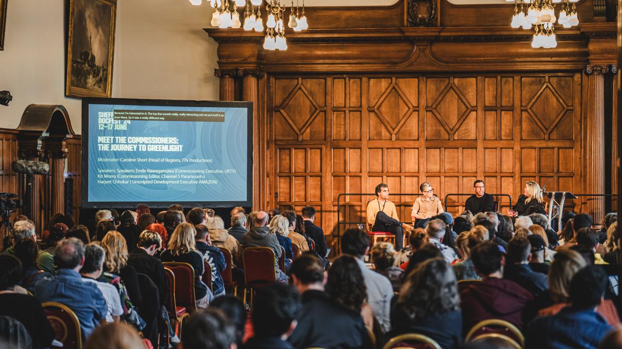 Busy room full of people sat on chairs, facing away from the camera. In the distance, a panel of people sit on a stage talking with microphones.