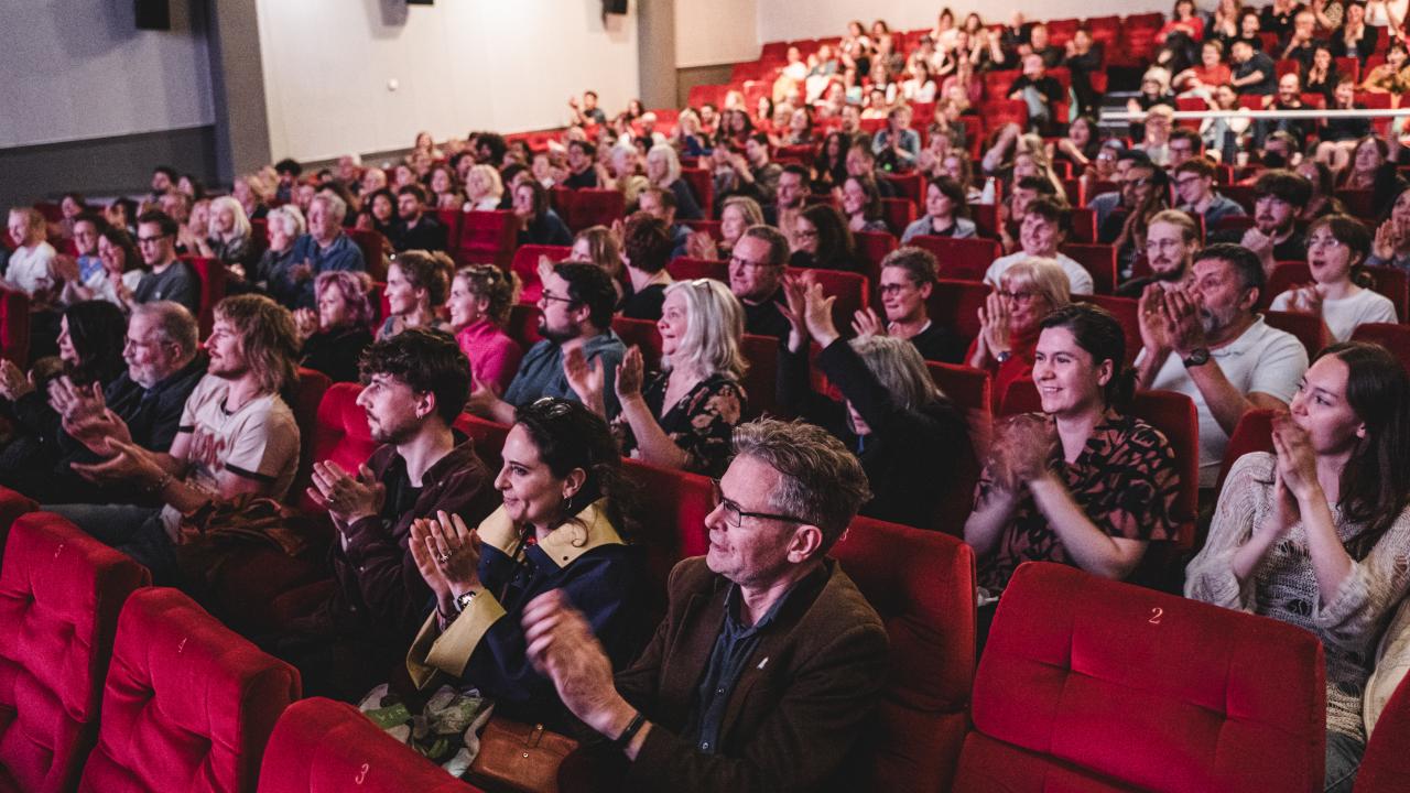 A happy crowd in red cinema seats applause with smiles on their faces.