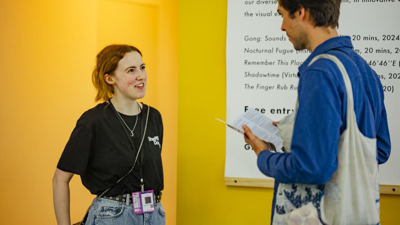 A bright yellow wall with two people standing in front of it. One, wearing a DocFest t-shirt smiles with their hands behind their back and the other, wearing a blue jacket, holds a piece of paper and looks forward.
